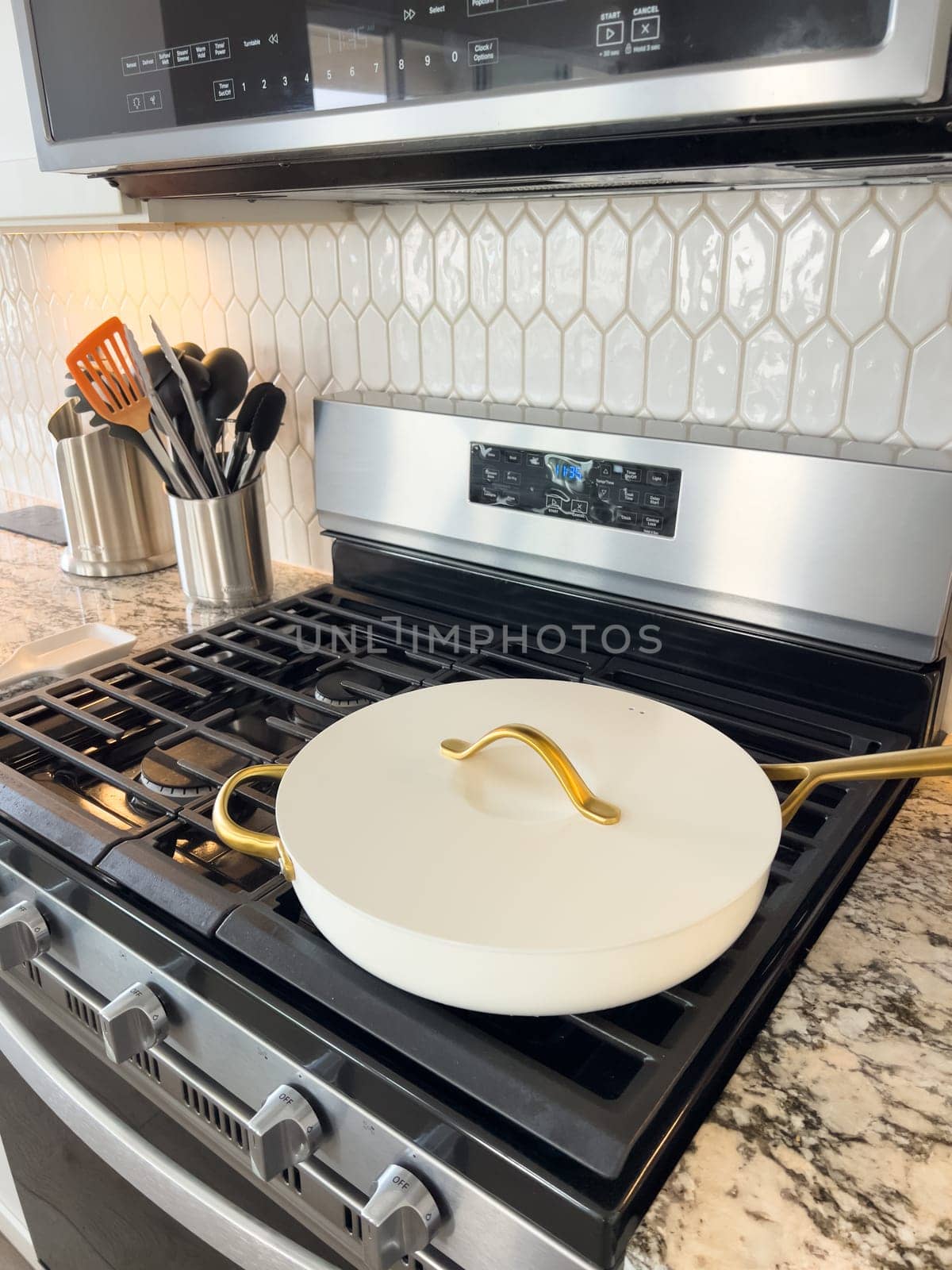 Castle Rock, Colorado, USA-January 17, 2024-A white ceramic pan with gold handles sits on a gas stove in a contemporary kitchen The stove features a stainless steel finish with multiple burners and control knobs A backsplash with white hexagonal tiles is visible in the background along with a utensil holder containing various kitchen tools and a bottle of cooking oil The scene showcases a clean and stylish cooking space