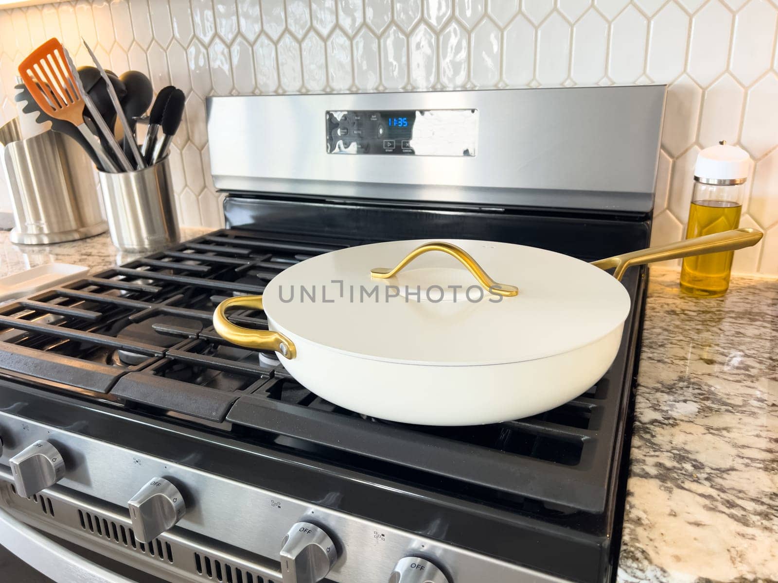 Castle Rock, Colorado, USA-January 17, 2024-A white ceramic pan with gold handles sits on a gas stove in a contemporary kitchen The stove features a stainless steel finish with multiple burners and control knobs A backsplash with white hexagonal tiles is visible in the background along with a utensil holder containing various kitchen tools and a bottle of cooking oil The scene showcases a clean and stylish cooking space