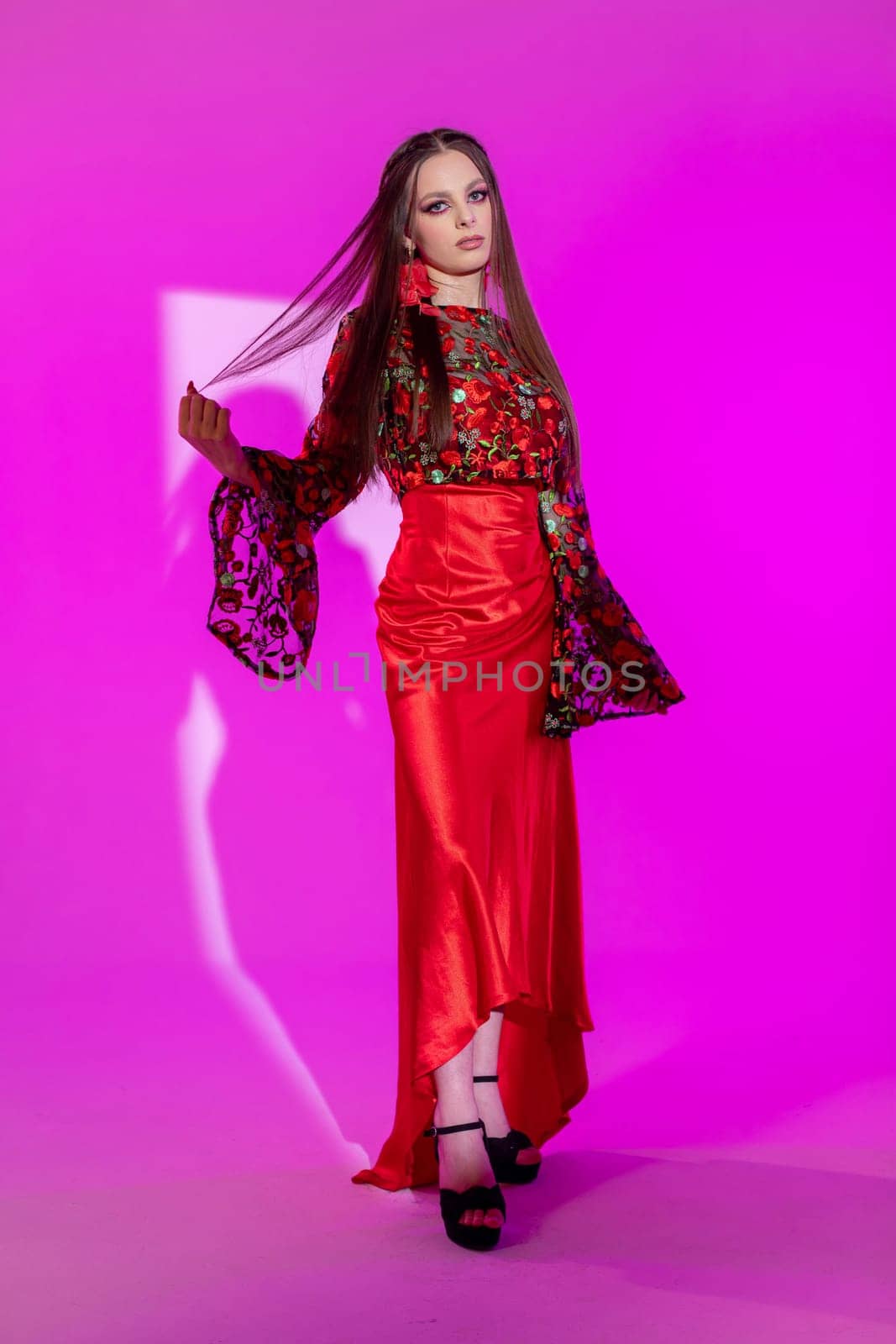 A stunning model poses in a bright pink setting, showcasing a striking red outfit with intricate details.