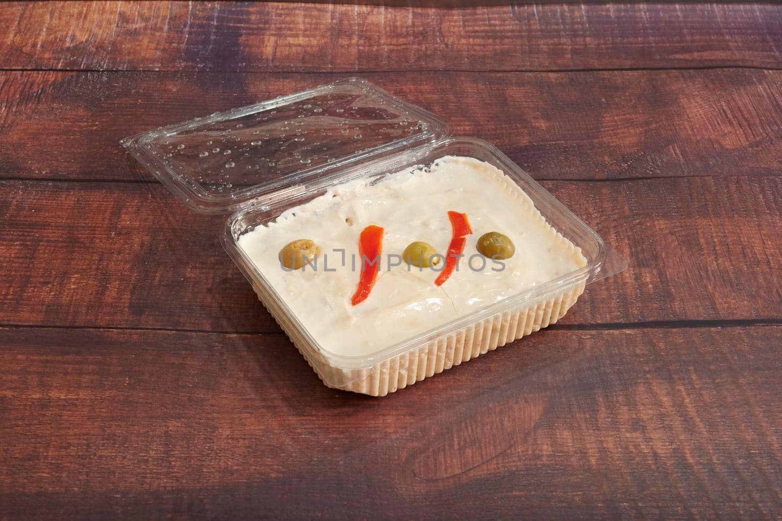 A small container of food with a lid on top. The food is a creamy white sauce with red peppers and olives