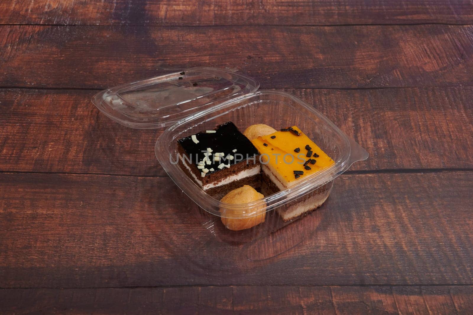 A box of assorted desserts sits on a wooden table. The desserts include a chocolate cake, a donut, and a piece of fruit. The box is open, revealing the delicious treats inside