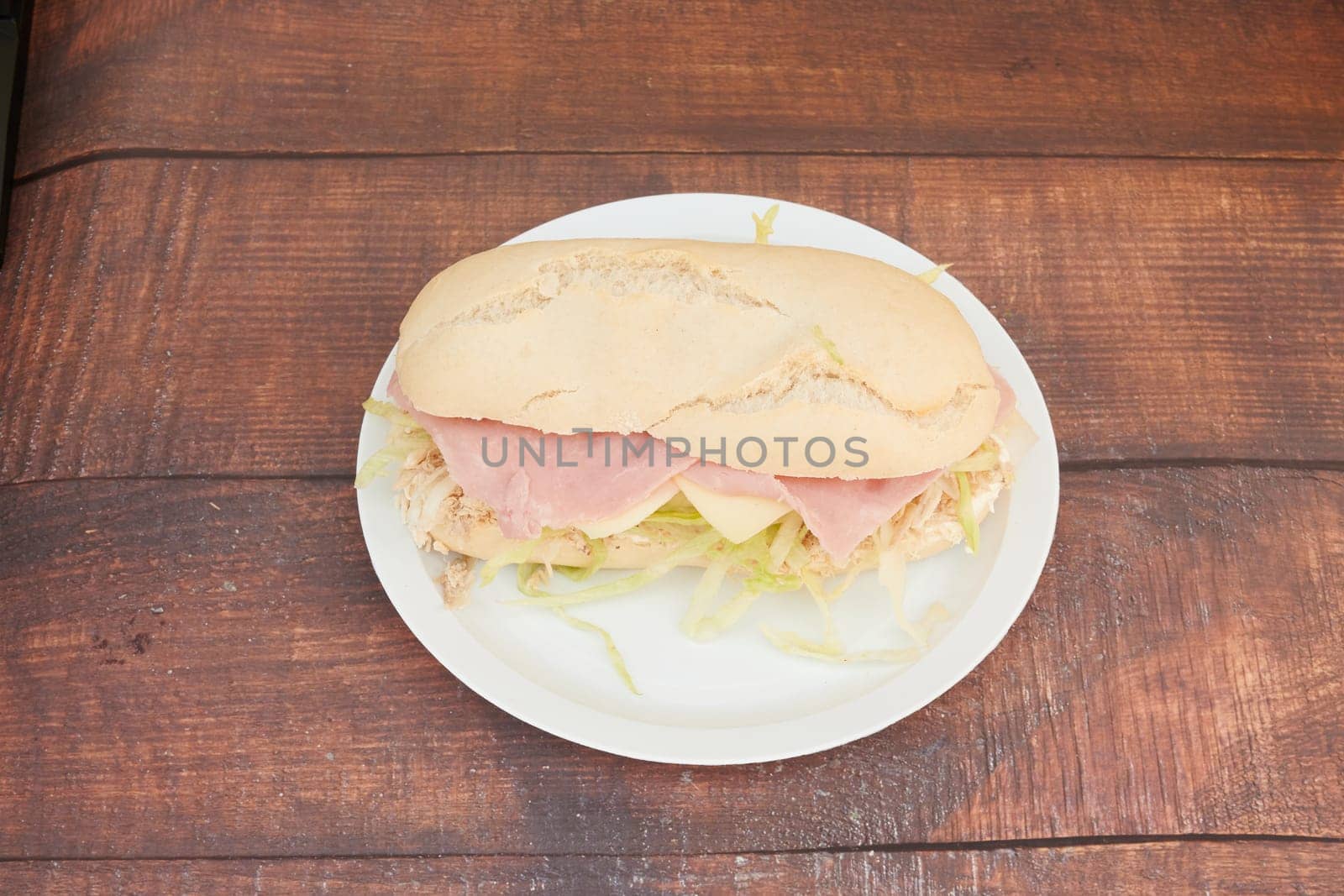 A sandwich is on a white plate on a wooden table. The sandwich is cut in half and has ham and lettuce on it