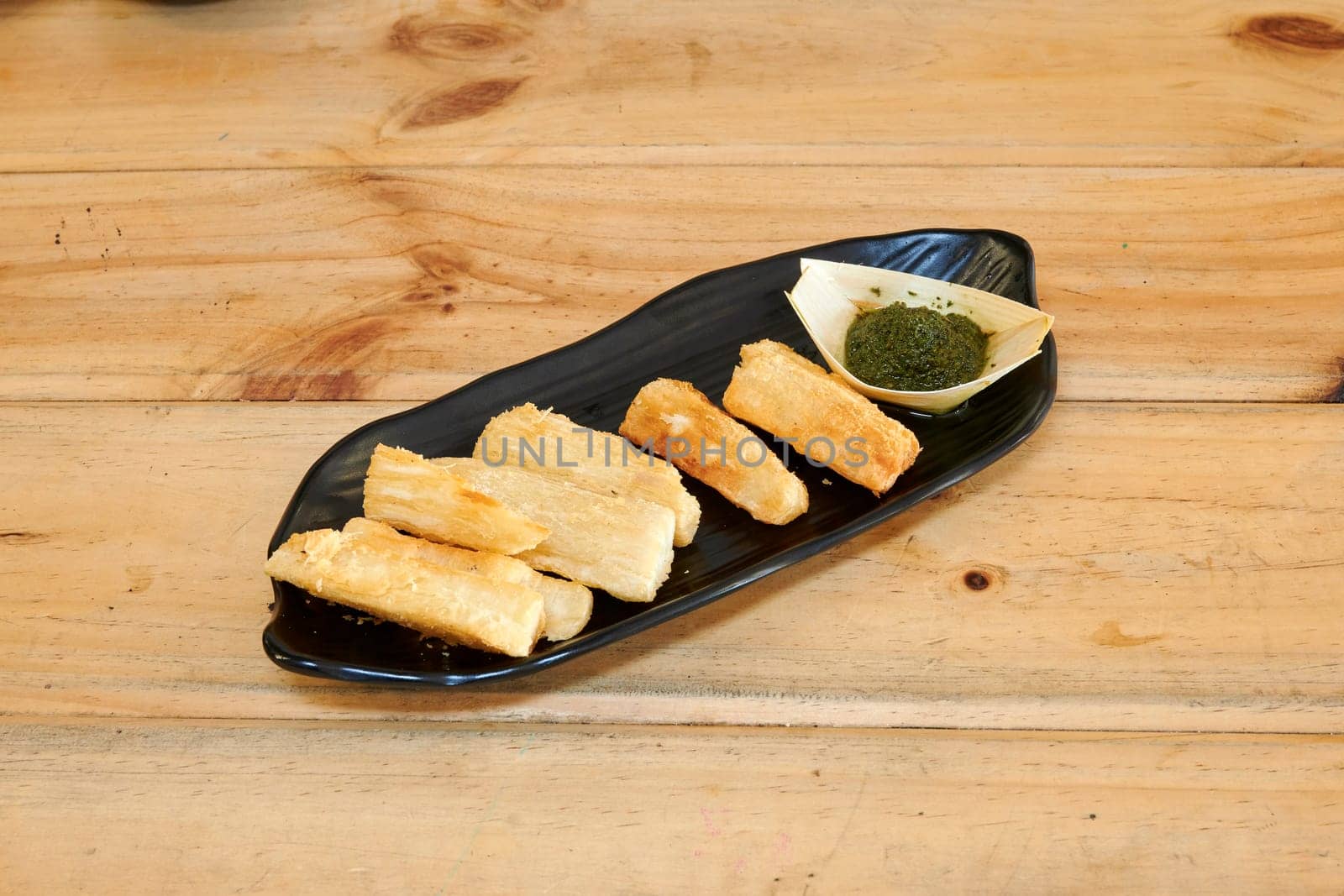 A plate of food with a green sauce on it. The food is cut into small pieces and arranged on a black plate. The plate is placed on a wooden table