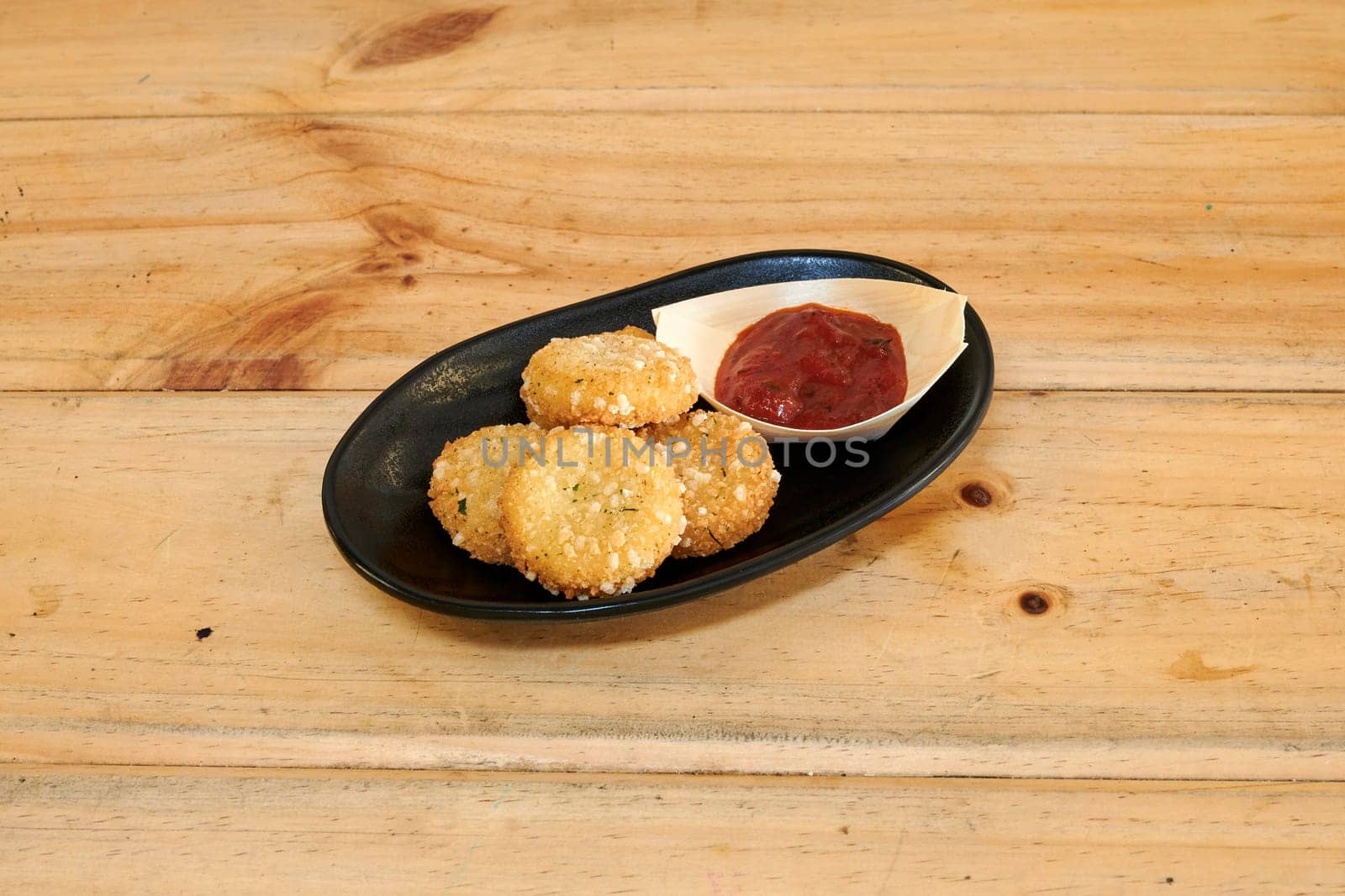 A plate of fried food with a red ketchup on a wooden table. The plate is black and the food is small