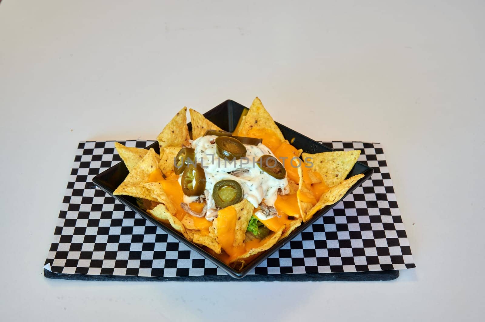 A plate of nachos with cheese and sour cream on top. The plate is on a checkered tablecloth