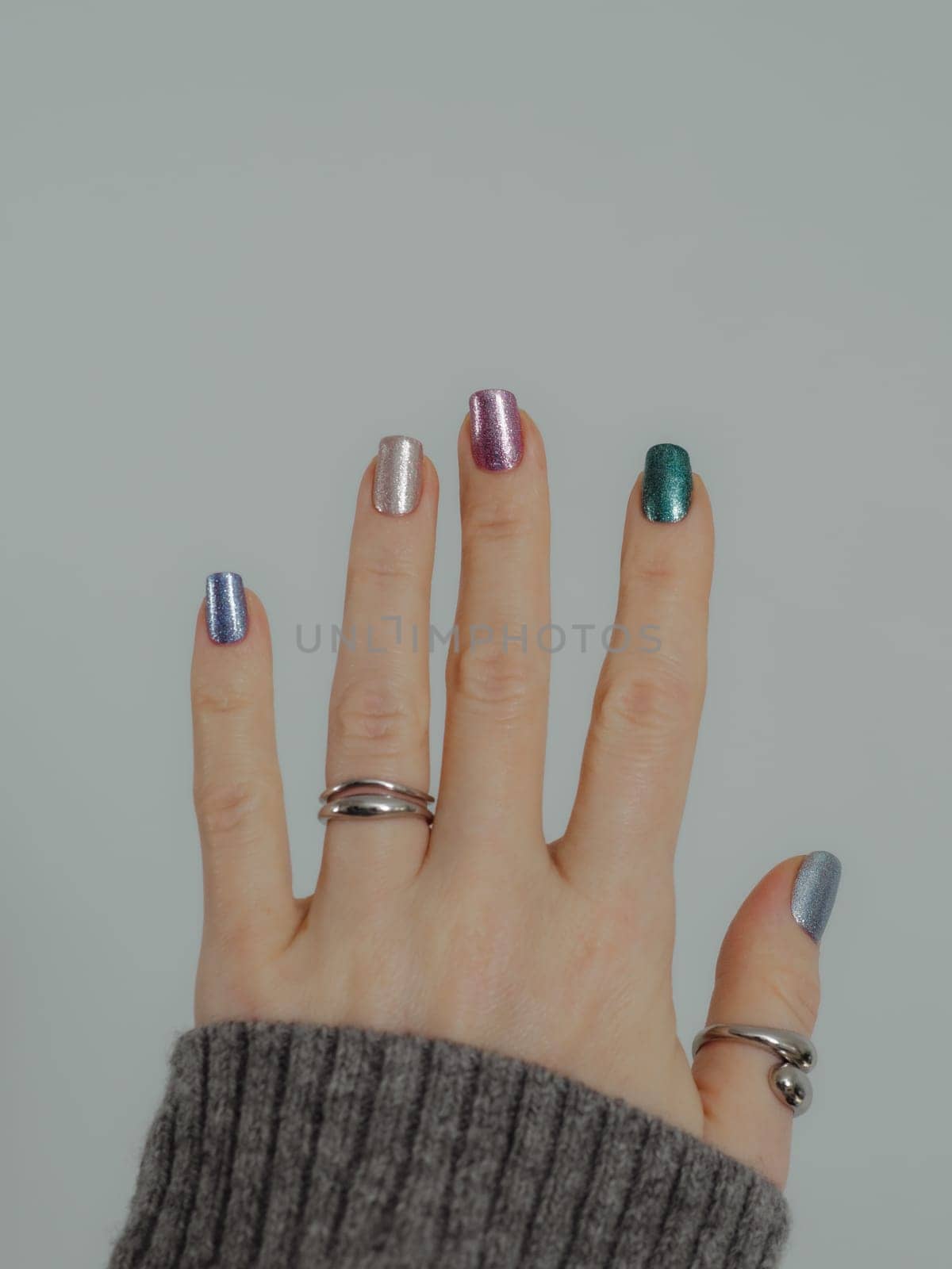 Skittles nails on hand. Colorful shiny manicure with holographic sparkle and metallic pigment. Female hand in gray sweater with multicolored nails on white wall background. Copy space. Vertical