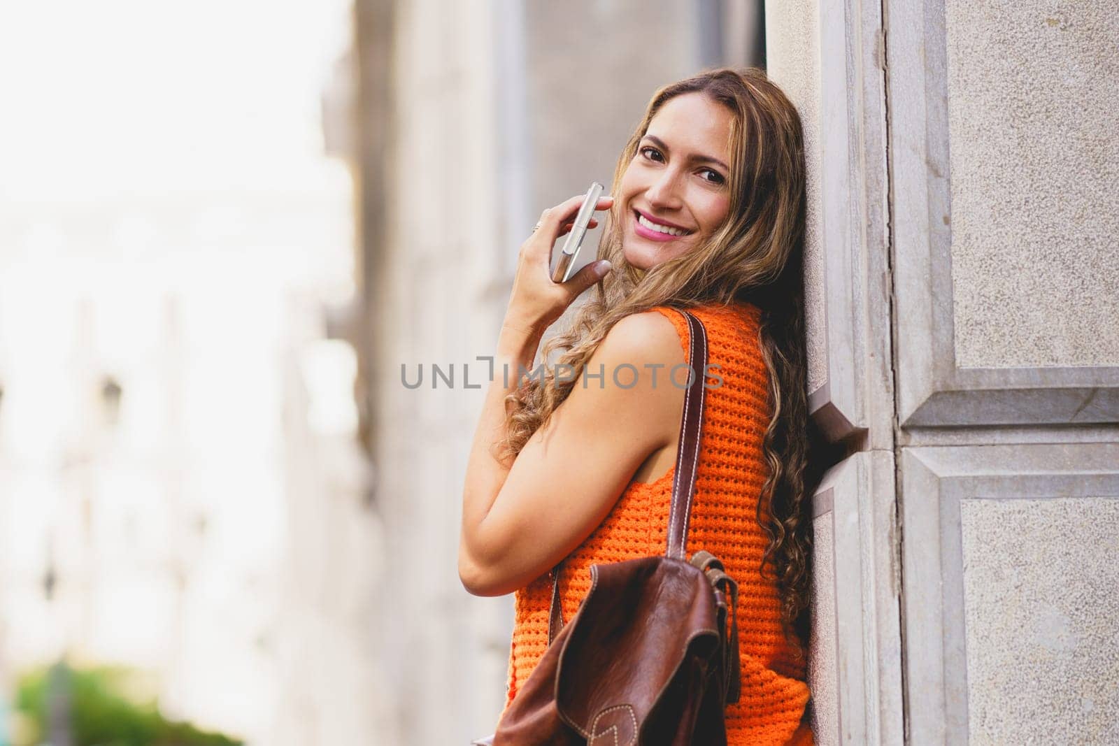 Latin woman leaning on an urban facade holding a mobile and smiling in the city