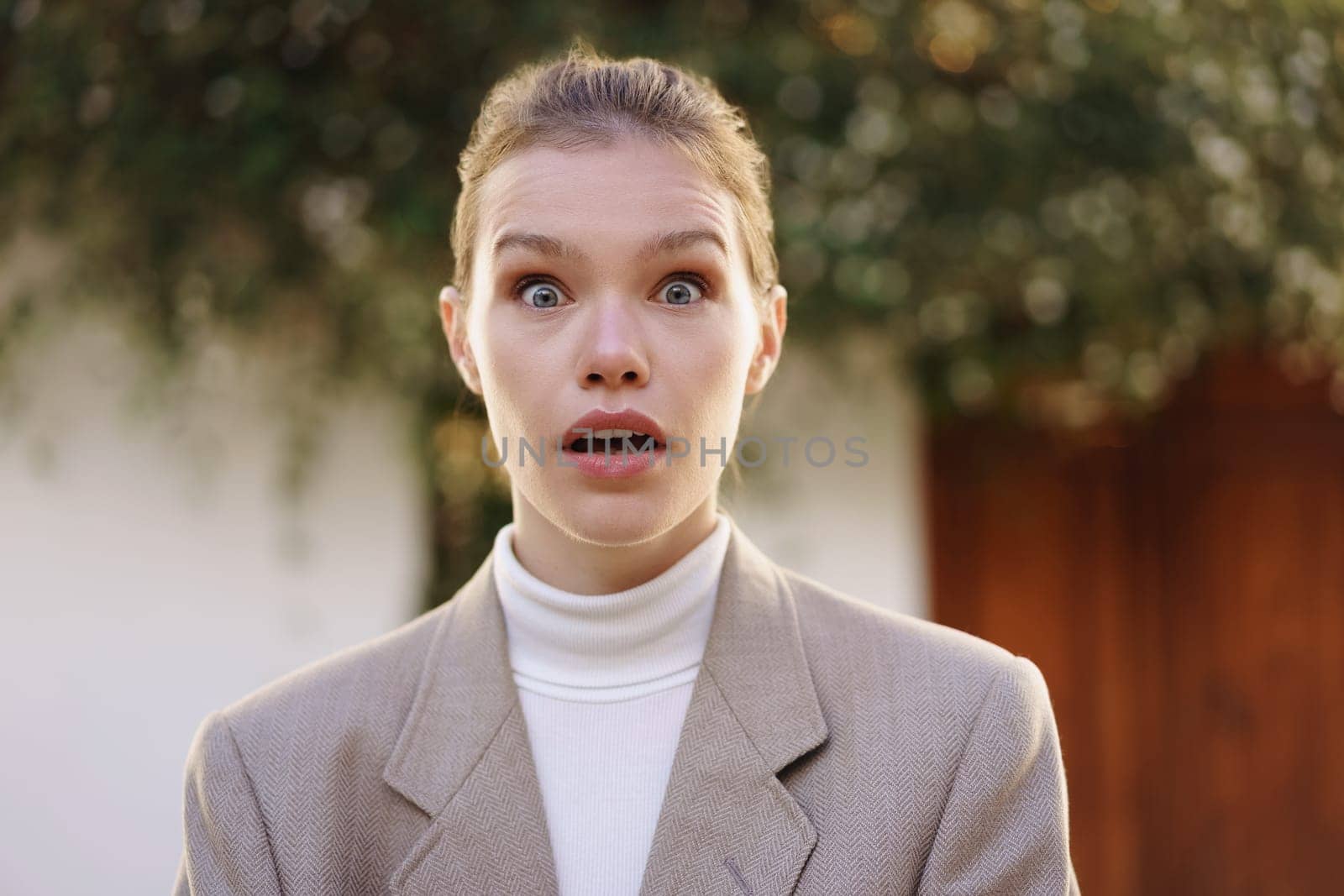 A woman is surprised while showcasing her chic blazer. This captivating image is framed with soft, blurred greenery, enhancing the dynamic expression and overall visual appeal, making it eyecatching