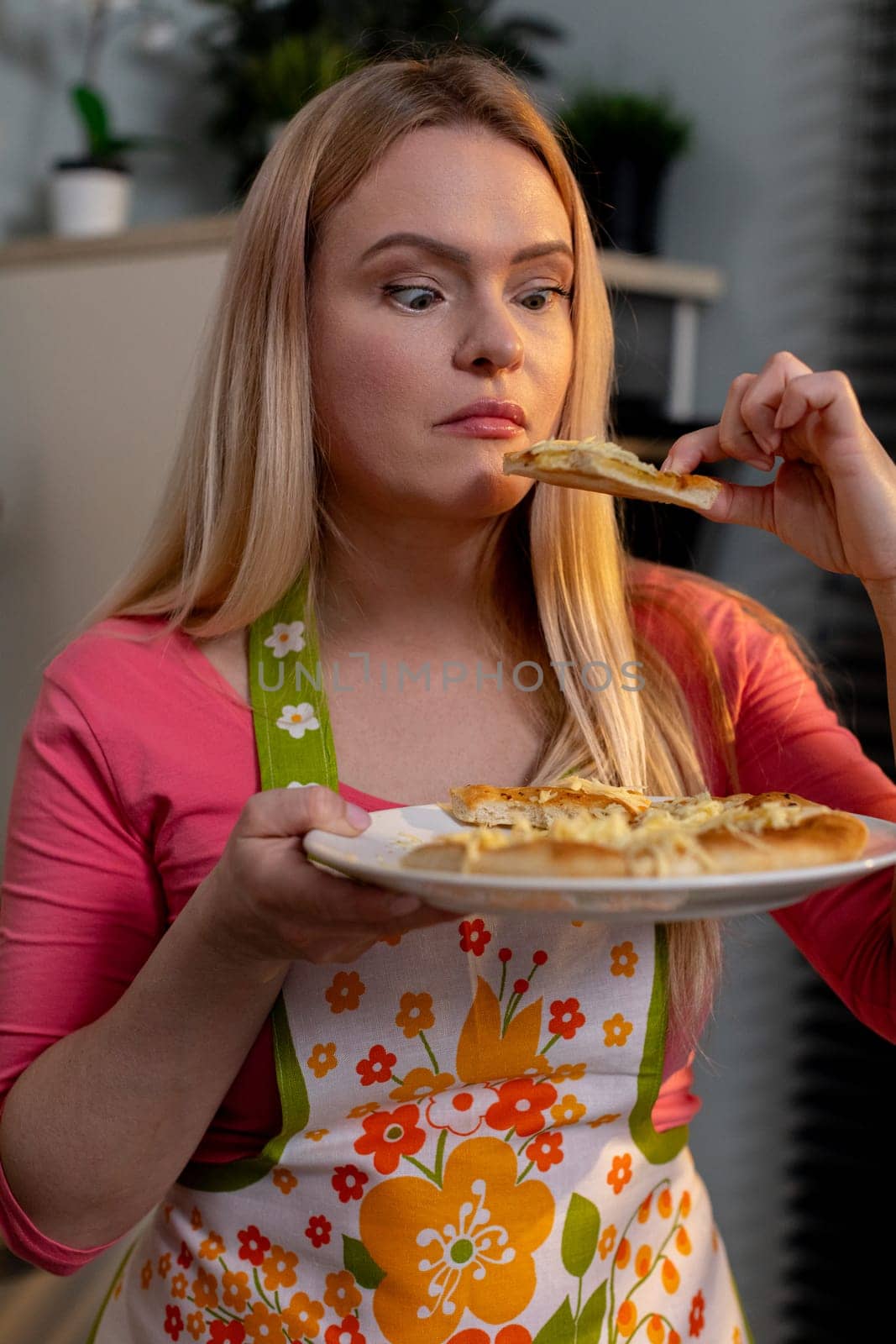 A happy woman is savoring a delicious slice of pizza in her vibrant kitchen while wearing a cheerful floral apron