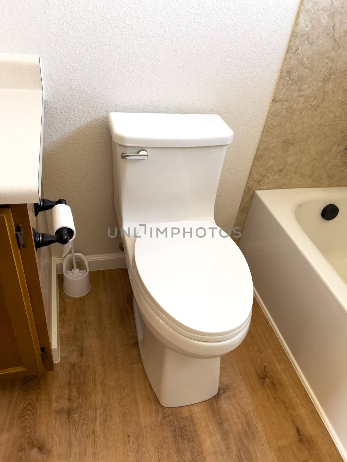 Broomfield, Colorado, USA-January 12, 2025-A clean white toilet with a closed lid is installed in a small bathroom with wood-style flooring The toilet is positioned against a white wall with a toilet paper holder mounted on the side and a small trash can nearby A bathtub is partially visible on the right side creating a compact yet functional bathroom space