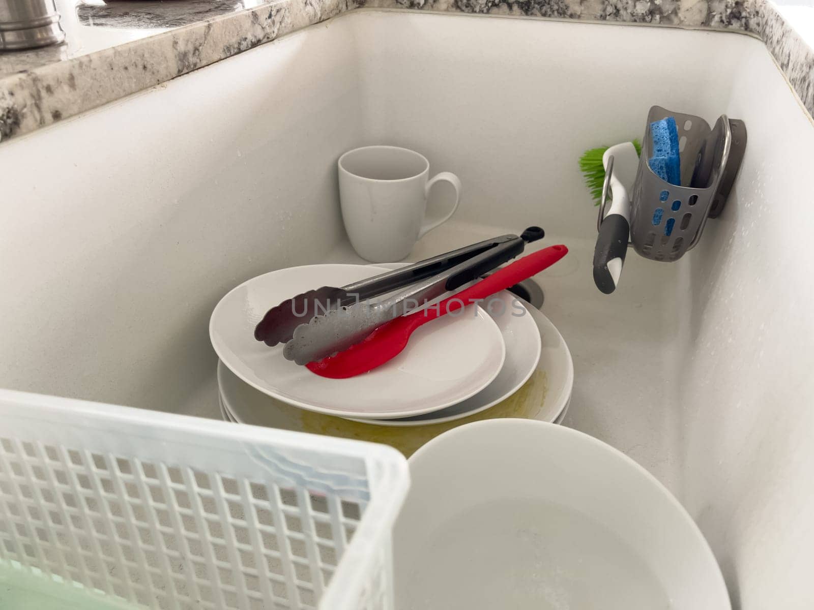 Castle Rock, Colorado, USA-January 18, 2024-A white kitchen sink filled with dirty dishes including plates bowls a coffee mug and utensils such as tongs and knives A dish rack with soapy water is placed on one side while a sponge and dish scrubber rest nearby The sink has a modern design with a garbage disposal drain