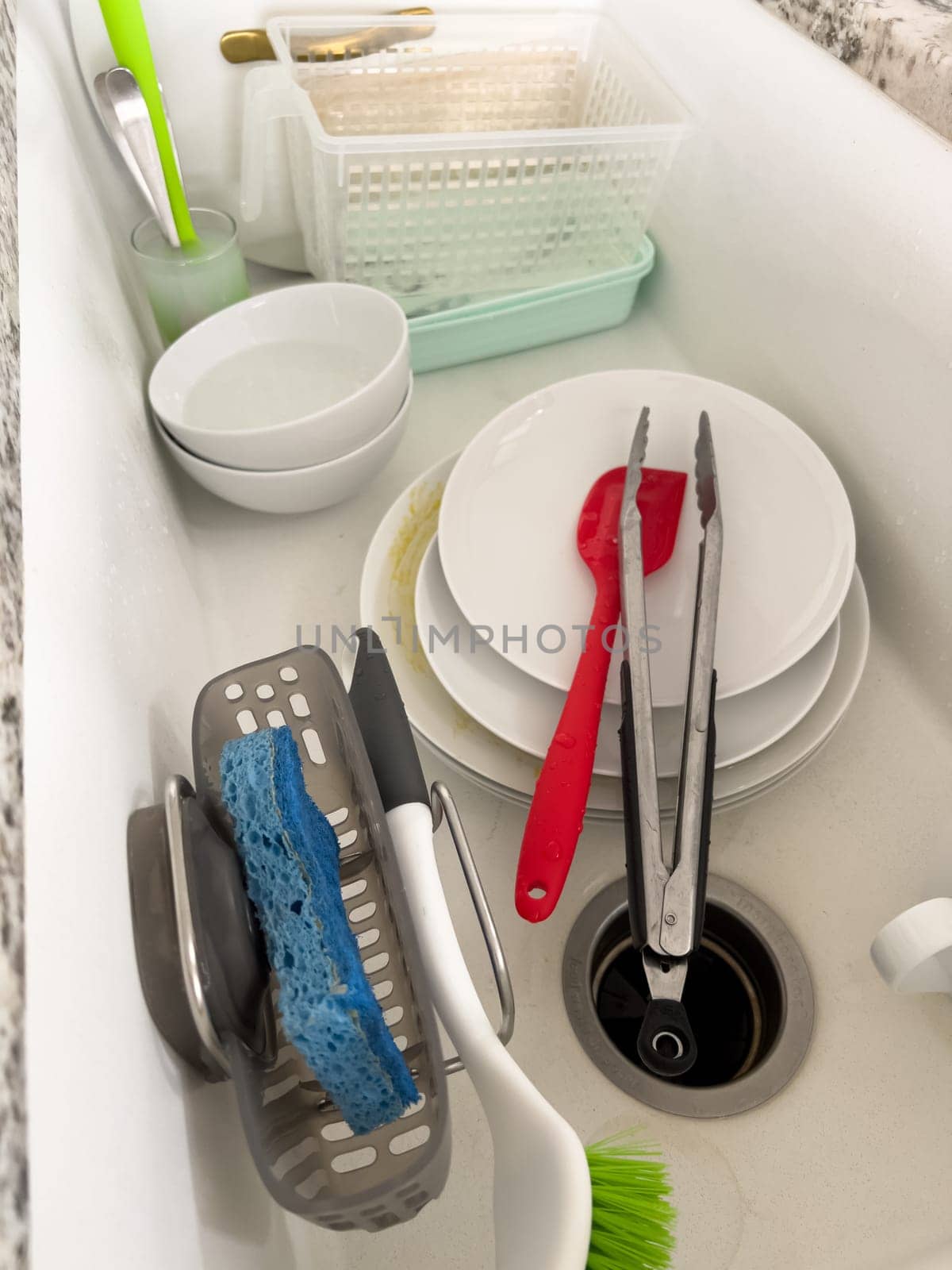 Castle Rock, Colorado, USA-January 18, 2024-A white kitchen sink filled with dirty dishes including plates bowls a coffee mug and utensils such as tongs and knives A dish rack with soapy water is placed on one side while a sponge and dish scrubber rest nearby The sink has a modern design with a garbage disposal drain
