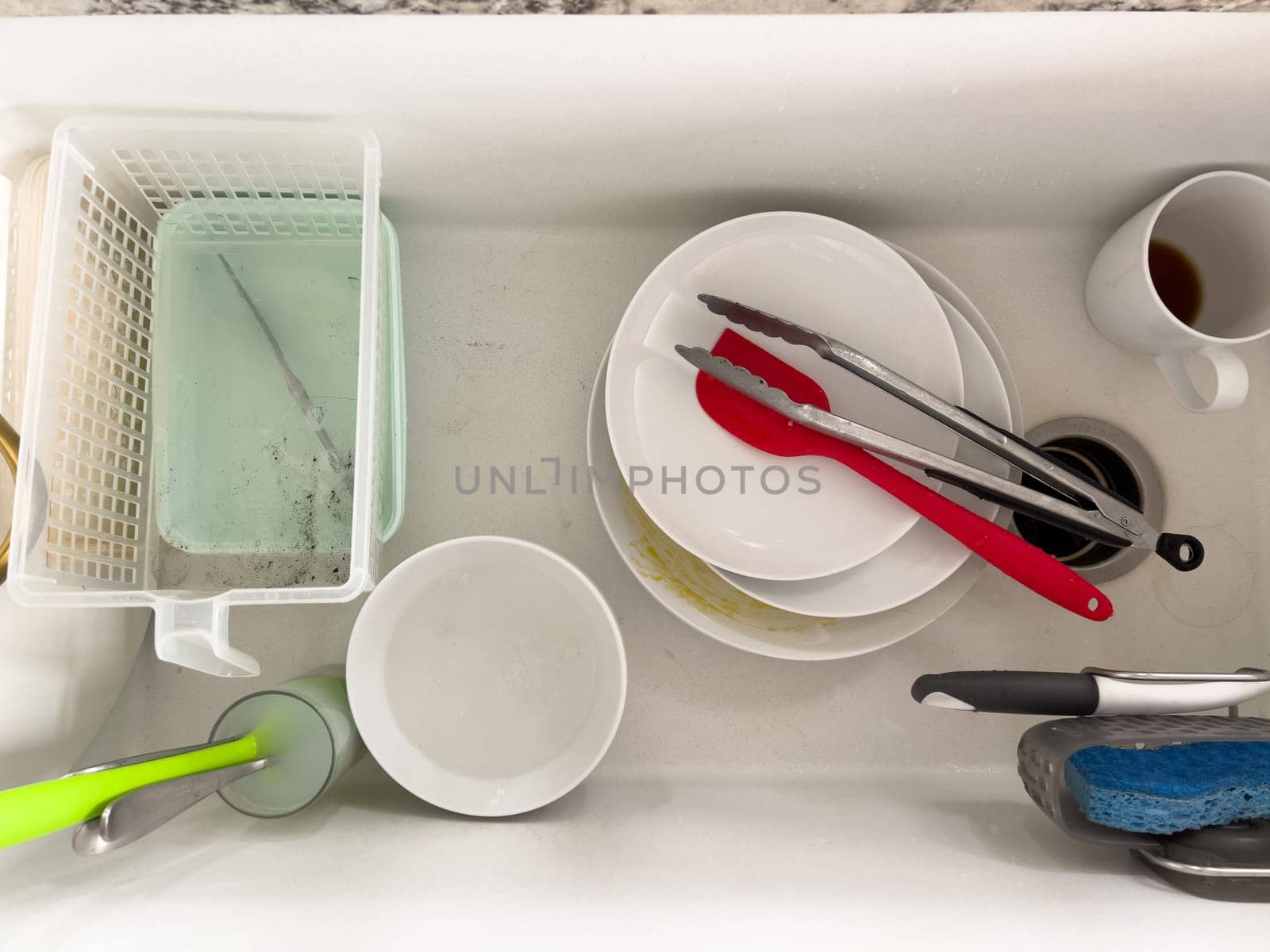 Castle Rock, Colorado, USA-January 18, 2024-A white kitchen sink filled with dirty dishes including plates bowls a coffee mug and utensils such as tongs and knives A dish rack with soapy water is placed on one side while a sponge and dish scrubber rest nearby The sink has a modern design with a garbage disposal drain