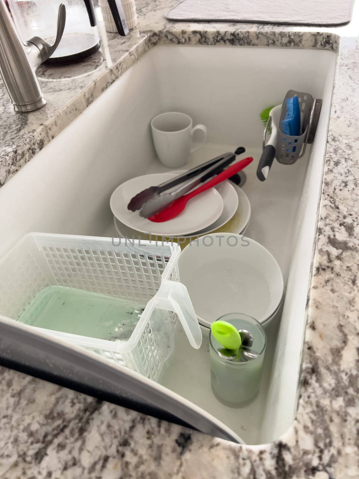 Castle Rock, Colorado, USA-January 18, 2024-A white kitchen sink filled with dirty dishes including plates bowls a coffee mug and utensils such as tongs and knives A dish rack with soapy water is placed on one side while a sponge and dish scrubber rest nearby The sink has a modern design with a garbage disposal drain