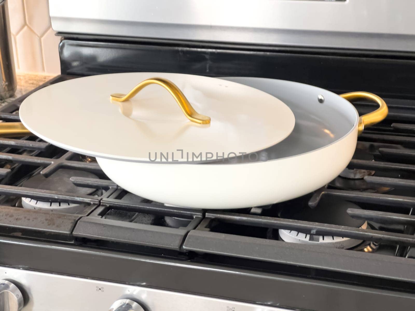Castle Rock, Colorado, USA-January 18, 2025-A white cooking pan with gold handles and a matching lid sits on a gas stove The pan has a sleek modern design with a nonstick surface The stainless steel stove has multiple burners and a digital display The kitchen counter features a marble design with various cooking utensils in the background