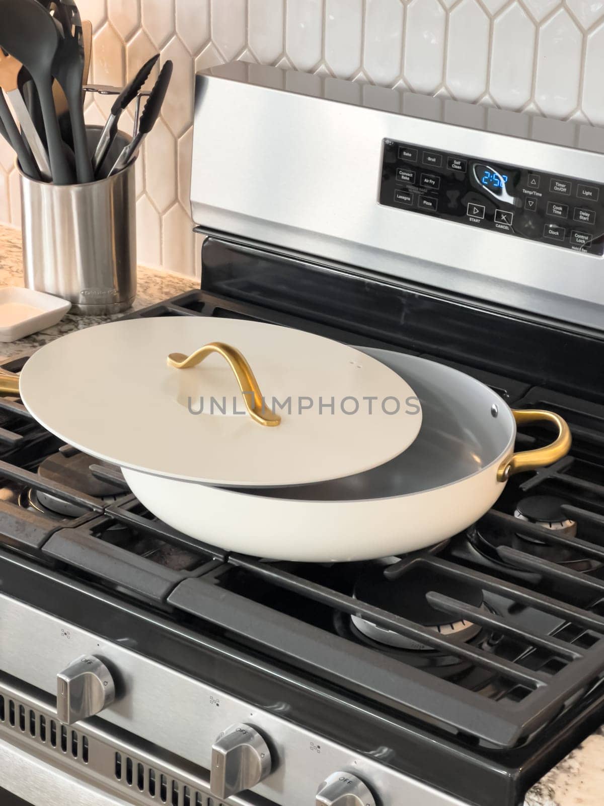 Castle Rock, Colorado, USA-January 18, 2025-A white cooking pan with gold handles and a matching lid sits on a gas stove The pan has a sleek modern design with a nonstick surface The stainless steel stove has multiple burners and a digital display The kitchen counter features a marble design with various cooking utensils in the background