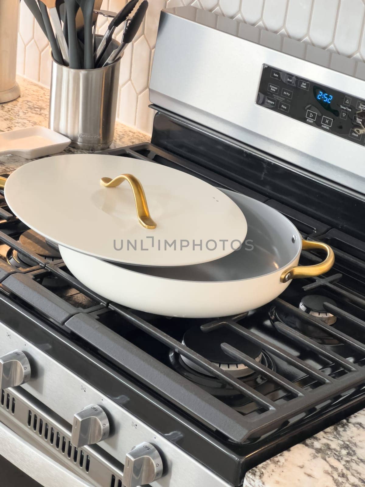 Castle Rock, Colorado, USA-January 18, 2025-A white cooking pan with gold handles and a matching lid sits on a gas stove The pan has a sleek modern design with a nonstick surface The stainless steel stove has multiple burners and a digital display The kitchen counter features a marble design with various cooking utensils in the background