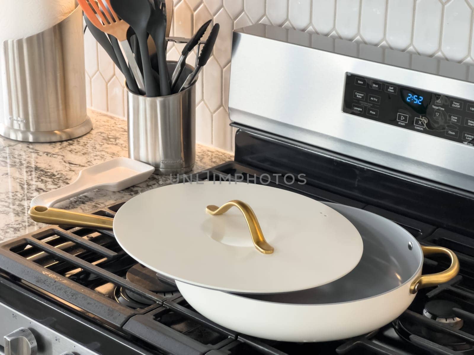 Castle Rock, Colorado, USA-January 18, 2025-A white cooking pan with gold handles and a matching lid sits on a gas stove The pan has a sleek modern design with a nonstick surface The stainless steel stove has multiple burners and a digital display The kitchen counter features a marble design with various cooking utensils in the background