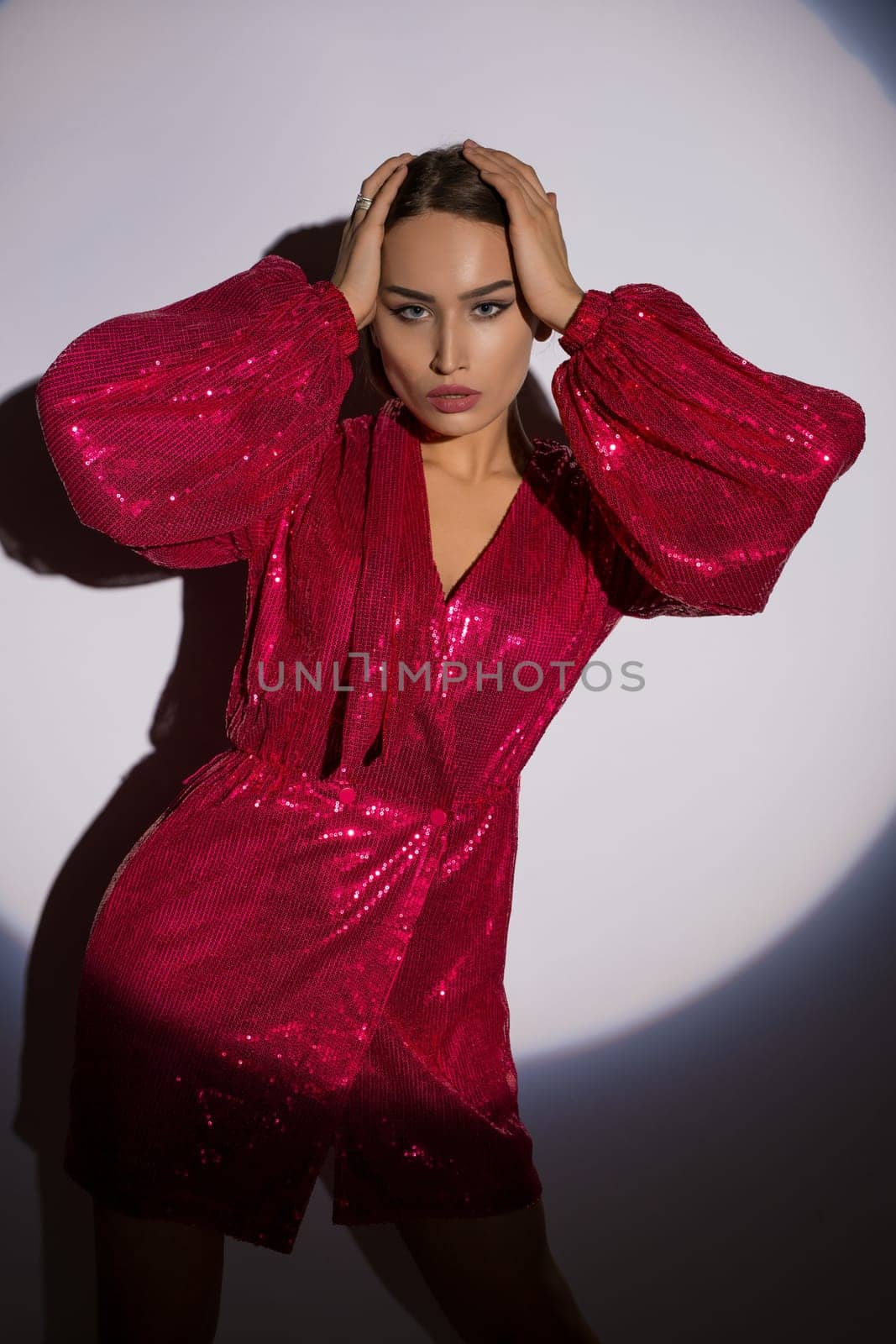 Confident female model wearing red glittering dress touching head while standing on white background in studio and looking at camera