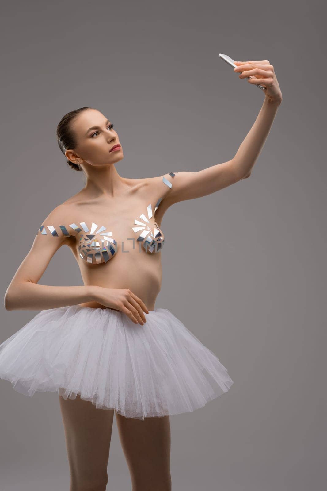 Attractive young slim female in ballet tutu skirt and with sticky tapes on breast taking selfie on smartphone against gray background