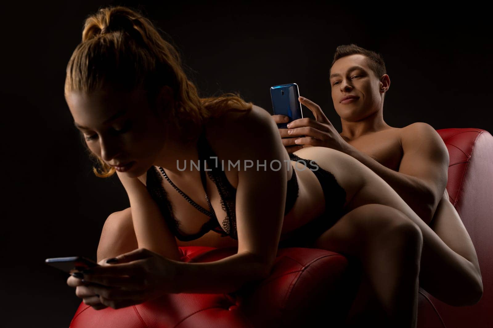 Young seductive woman in sexy lingerie and handsome brutal boyfriend browsing mobile phones while relaxing on red leather chair in dark studio