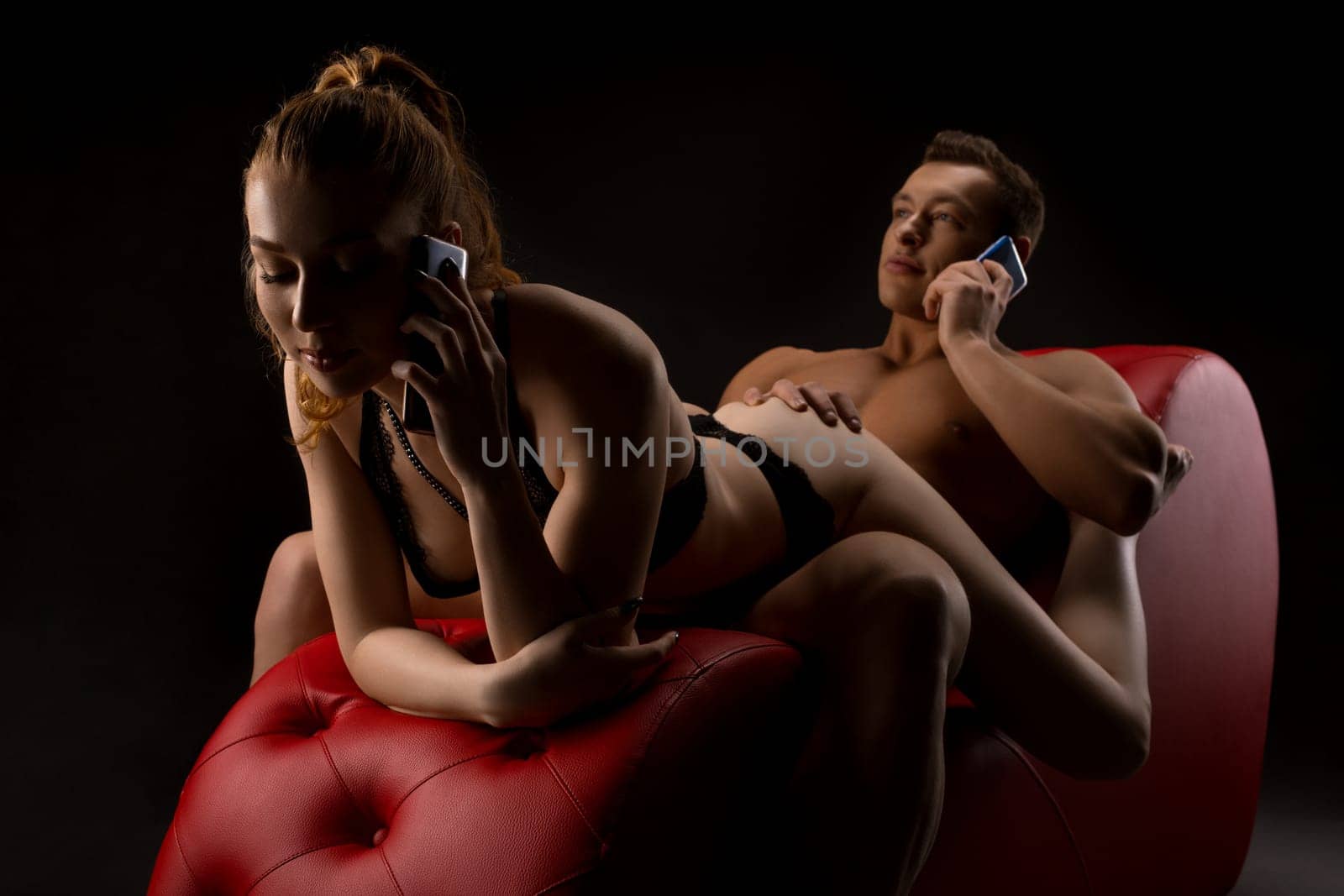 Young seductive woman in sexy lingerie and handsome brutal boyfriend call mobile phones while relaxing on red leather chair in dark studio