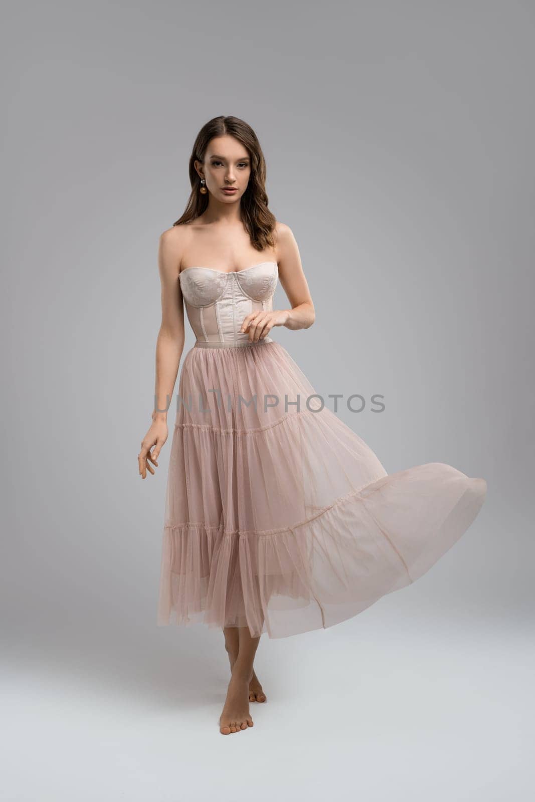 Full body of charming tender young female model wearing white corset and tulle skirt looking at camera while standing in light studio