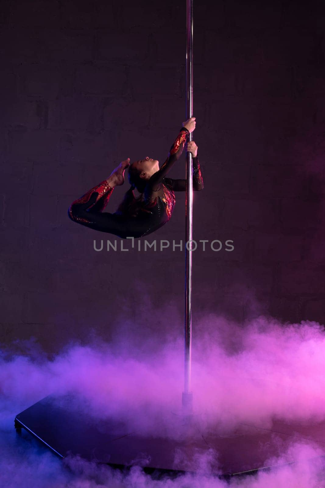 Full body side view of flexible teen female dancer doing aerial trick on pylon during performance on stage with smoke and neon illumination