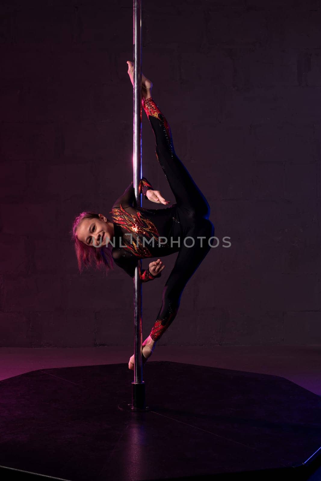 Full body of flexible teen female gymnast in leotard performing acrobatic standing splits near pole in dark studio