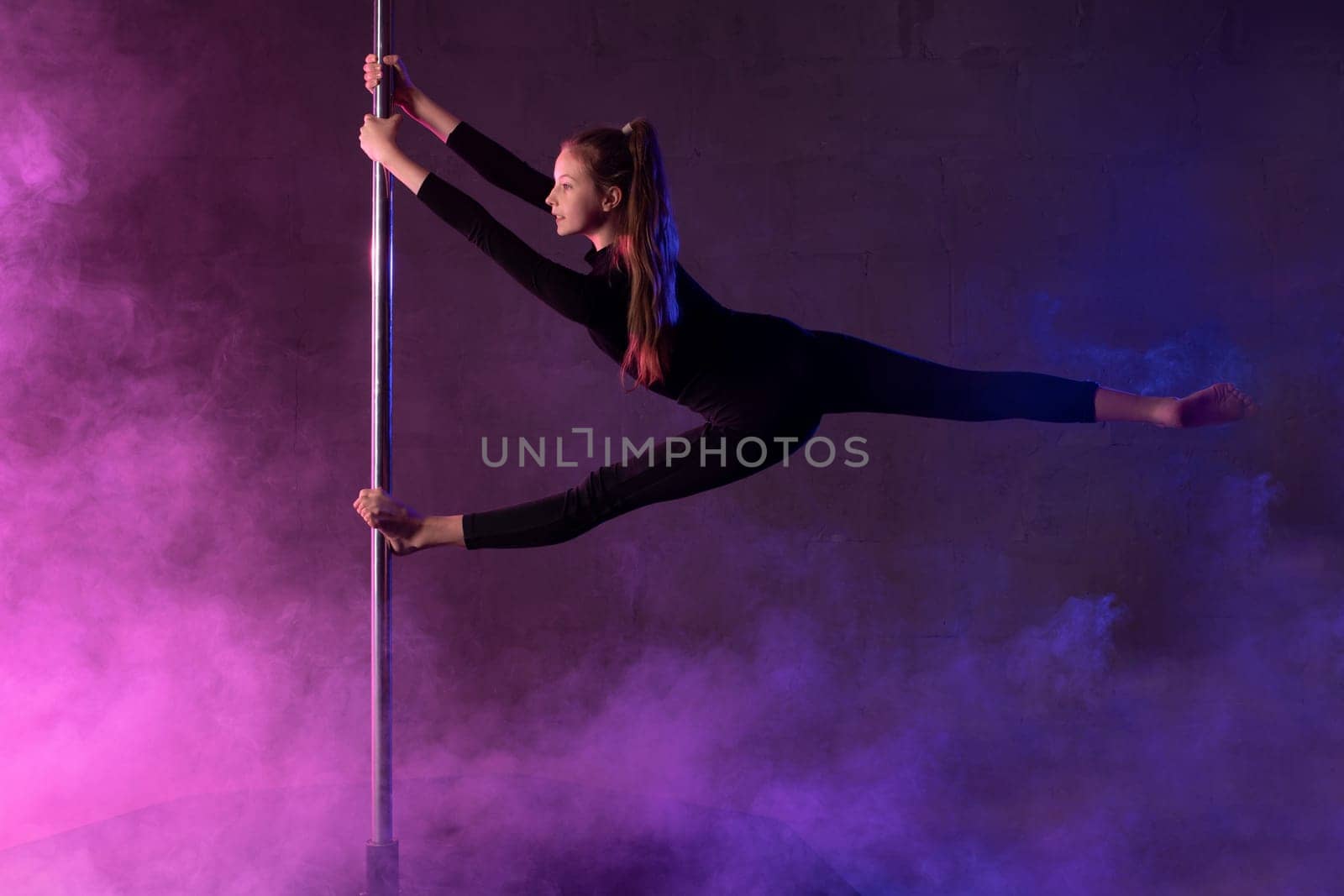 Full body side view of young flexible teen female dancer in black hanging on pole and doing splits during performance under neon light and smoke