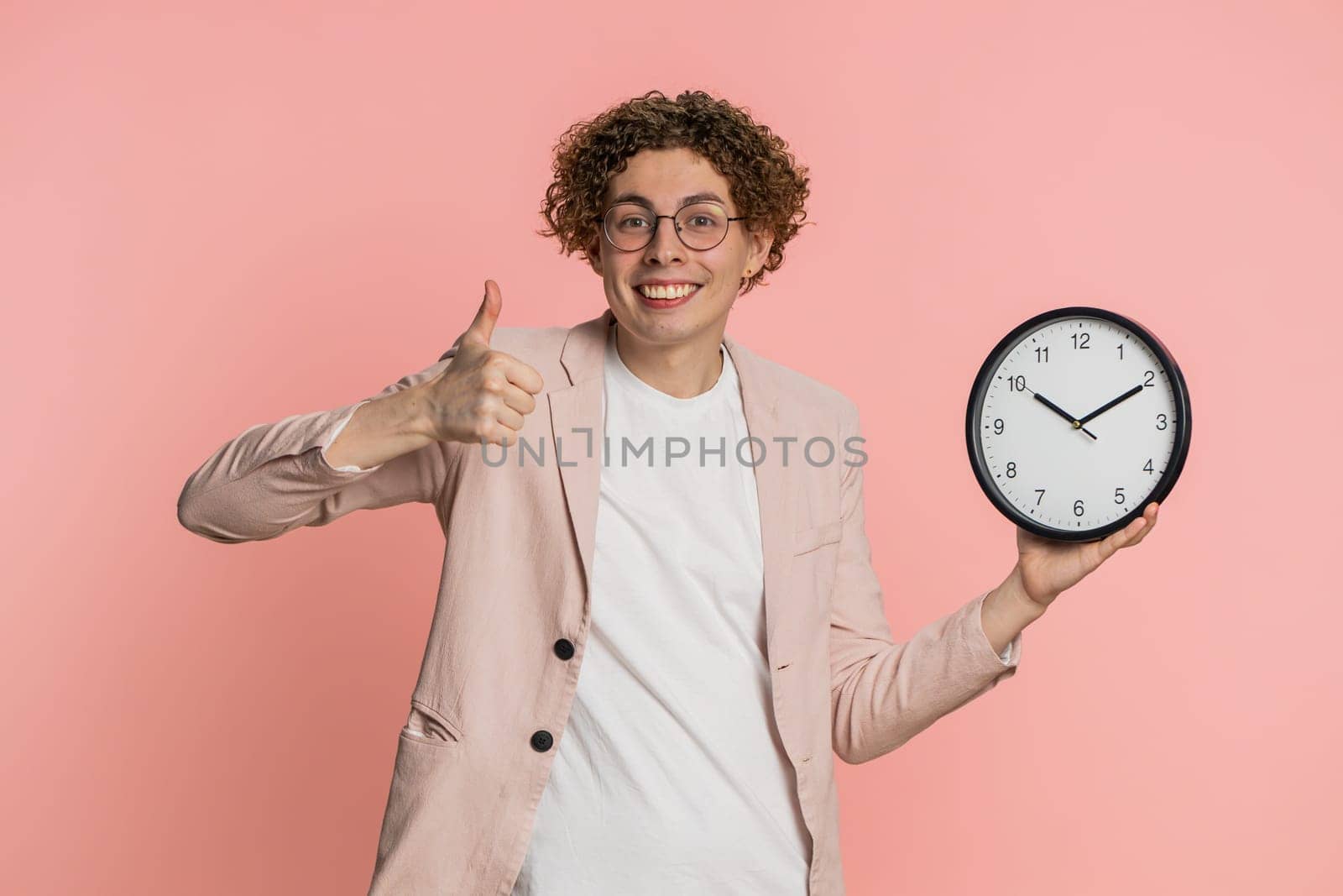 It is your time, hurry up. Caucasian man showing time on wall office clock, ok thumb up, approve, pointing finger at camera, advertisement, deadline. Curly haired young guy isolated on pink background