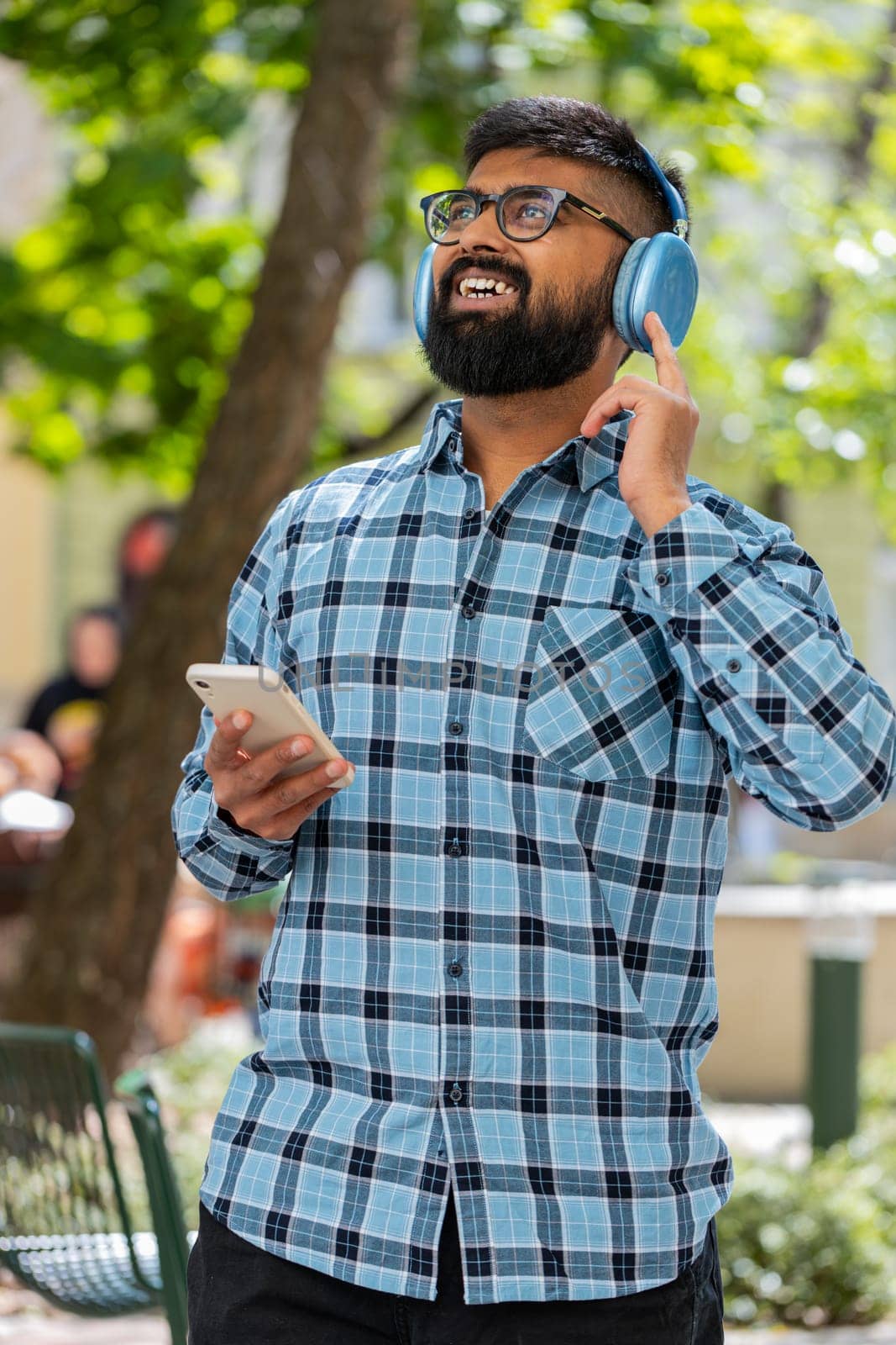 Happy overjoyed Indian bearded man dancing in wireless headphones listening favorite energetic music, celebrating victory win outdoors. Hispanic guy on urban city street. Town lifestyles. Vertical