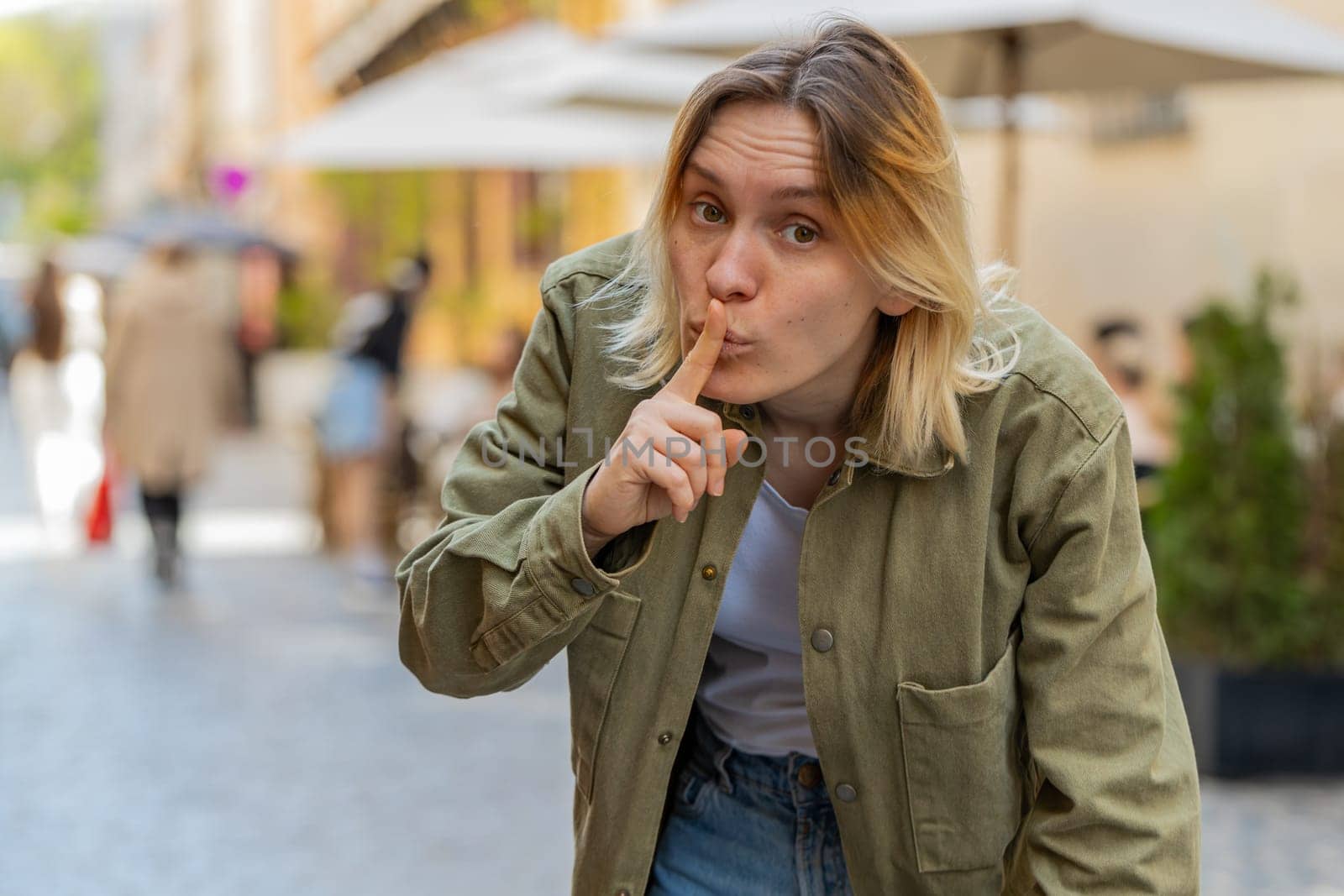 Shh be quiet please. Young woman presses index finger to lips makes silence hush gesture sign do not tells gossip secret outdoors. Caucasian girl tourist standing on urban city street. Town lifestyles