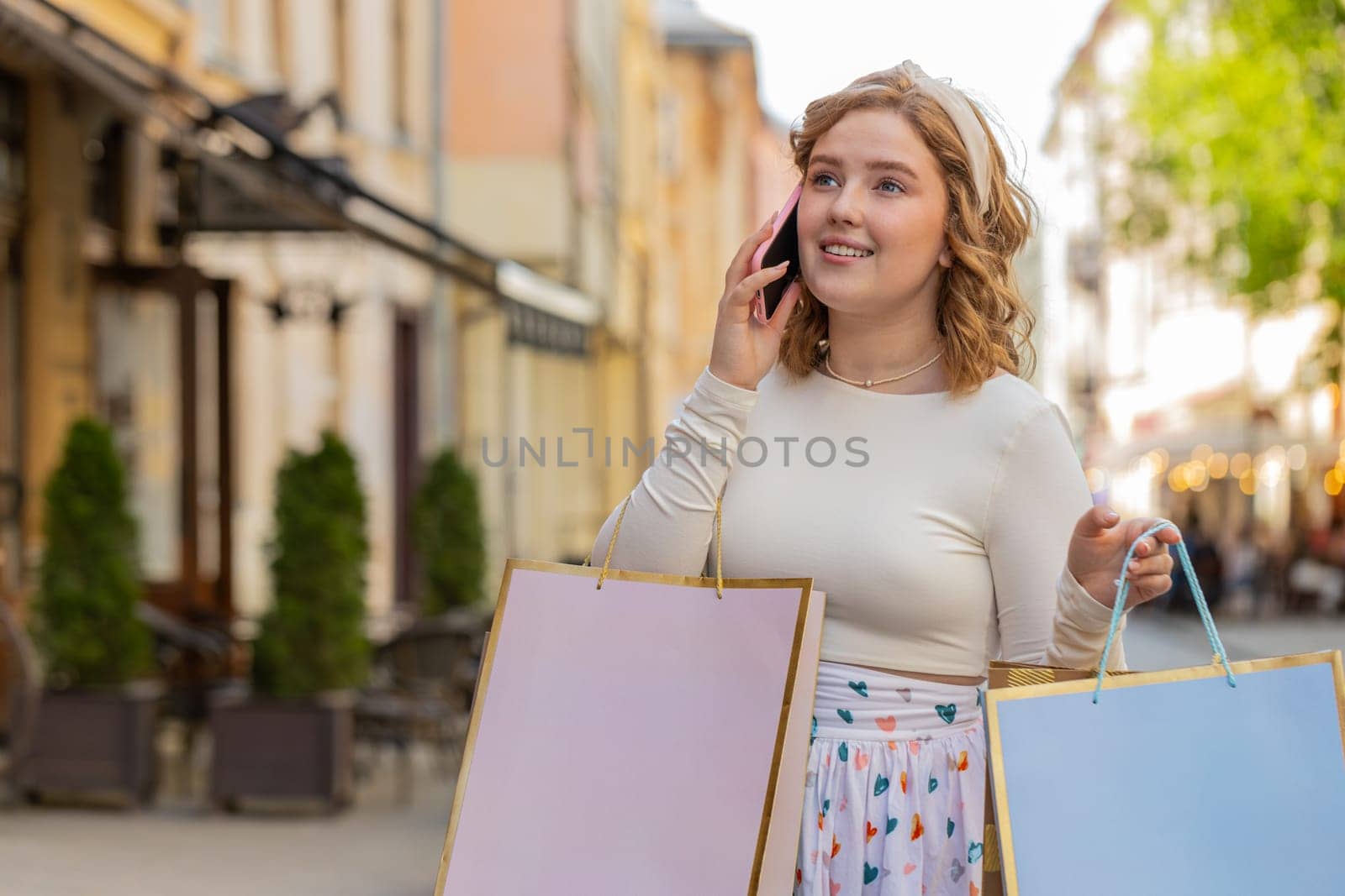 Happy smiling red hair woman shopaholic consumer after shopping sale with full gifts bags outdoors. Girl tourist traveler walking along city street talking on smartphone mobile remote conversation.