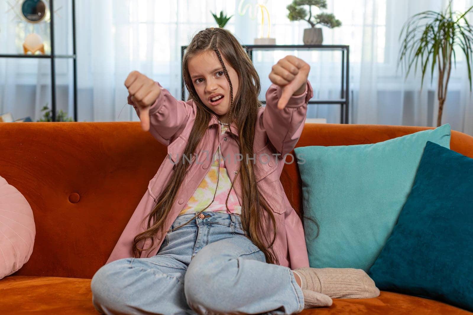 Dislike. Upset girl child showing thumbs down sign gesture, expressing discontent, disapproval, dissatisfied bad work at modern home apartment indoors. Displeased teenager kid in living room on sofa.