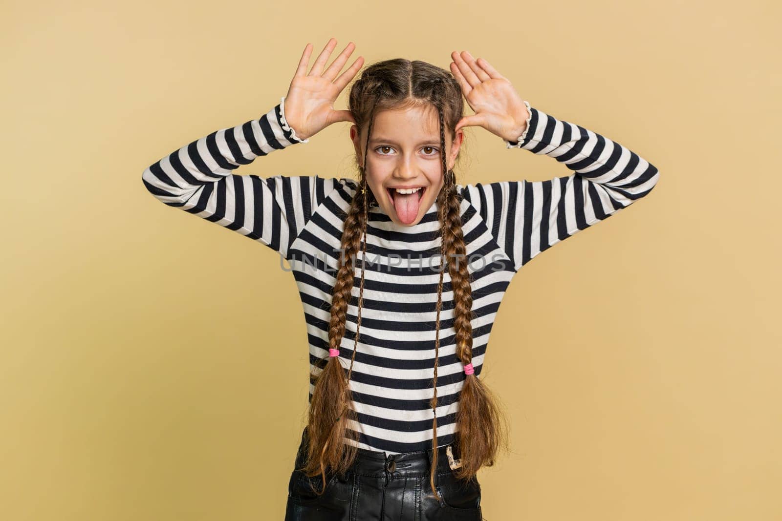 Cheerful funny young school girl showing tongue making faces at camera, fooling around, joking, aping with silly face teasing bullying, abuse. Preteen female child kid 10 years old on beige background