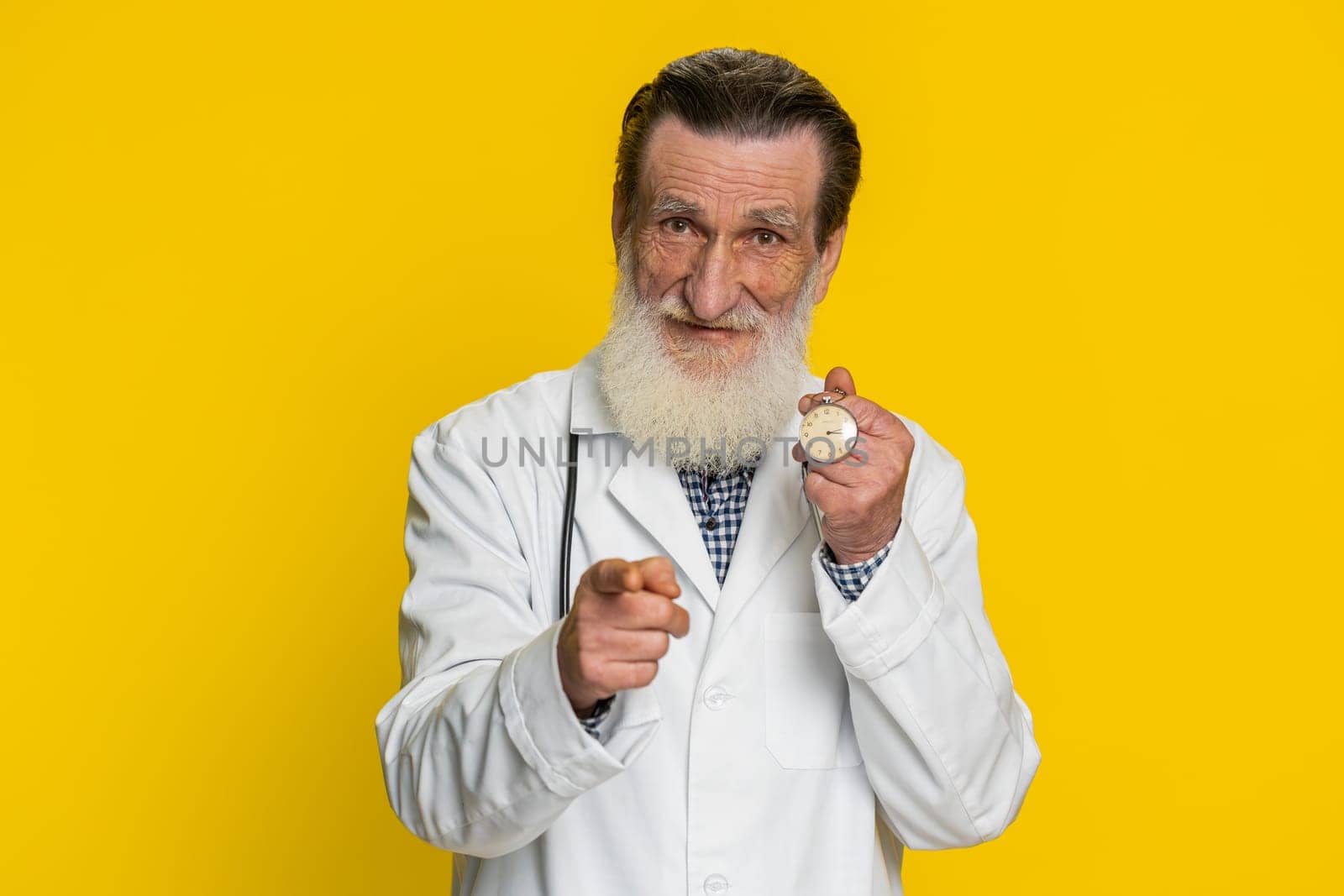 It is your time to heal. Senior mature doctor cardiologist man showing time on pocket watch clock ok thumb up approve pointing finger at camera. Apothecary pharmacy grandfather on yellow background