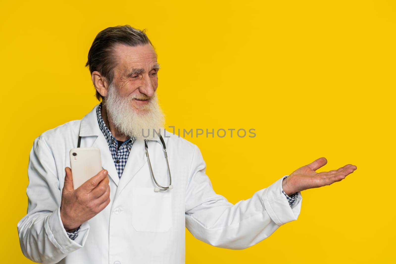 Senior doctor cardiologist man using smartphone showing pointing empty place, advertising application area, positive feedback, copy-space. Apothecary pharmacy grandfather isolated on yellow background