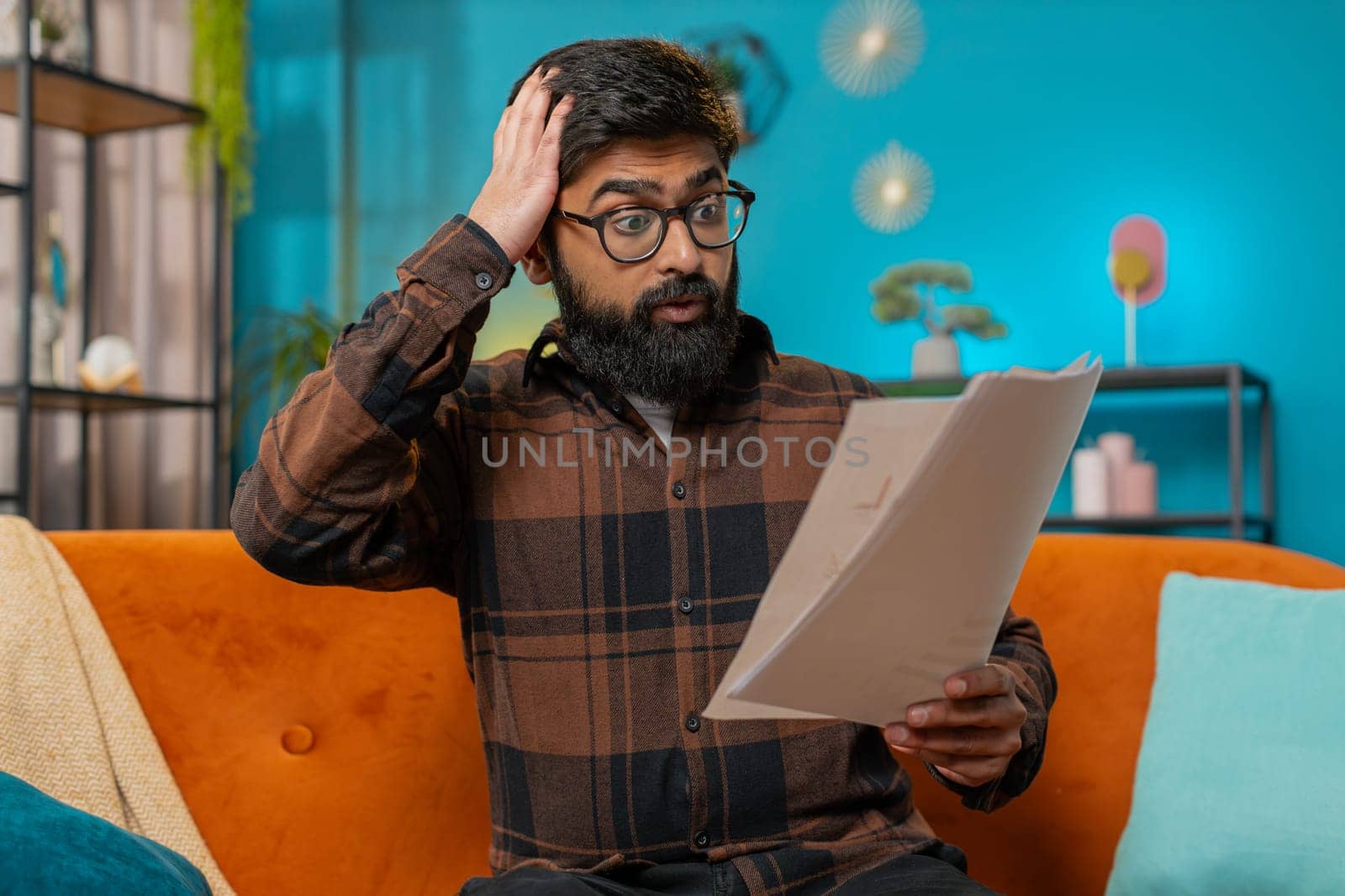 Frustrated upset amazed Indian business man works at home sofa looking at bills paperwork reports documents gets angry, frustrated, stress bankruptcy financial problem. Stresses freelancer at home