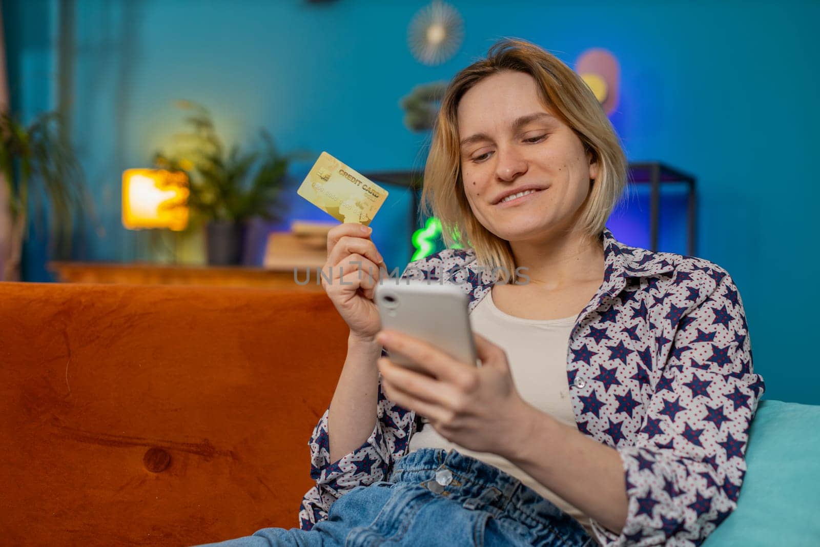 Happy young woman doing payments online purchase shopping in internet store. Smiling Caucasian girl holding banking credit card involved in verification process in mobile application on sofa at home.