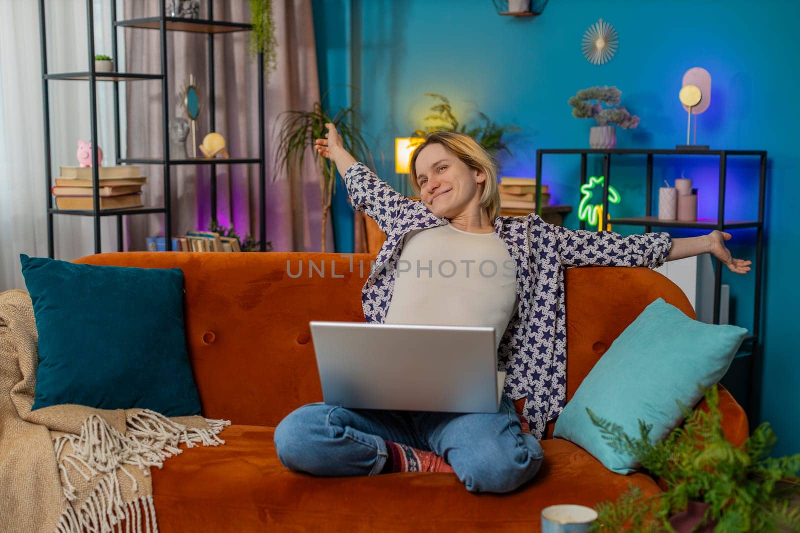 Happy young relaxed blonde woman sitting on couch after finishing laptop work in living room. Caucasian girl student working online, remote job at home sofa. E-learning, browsing internet on netbook.