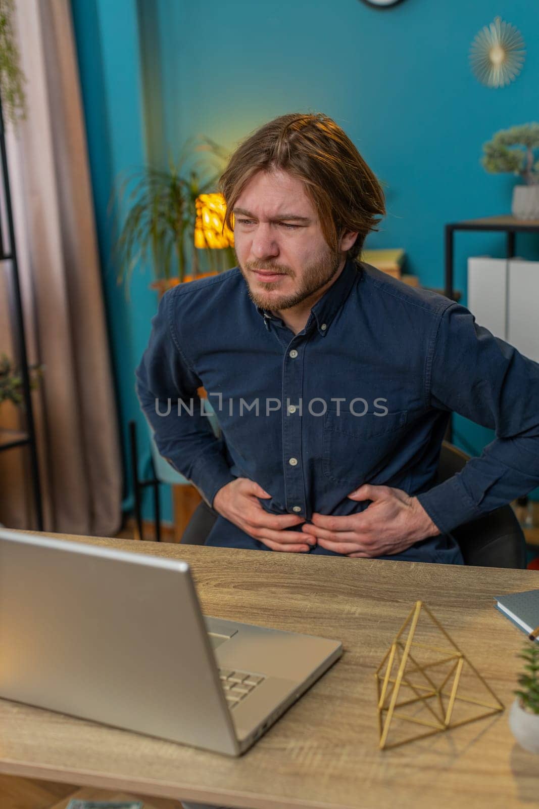 Disappointed freelancer feeling overeating during work process on laptop in home office. Upset businessman freelancer guy man touching stomach and experiencing discomfort and sharp pain. Vertical