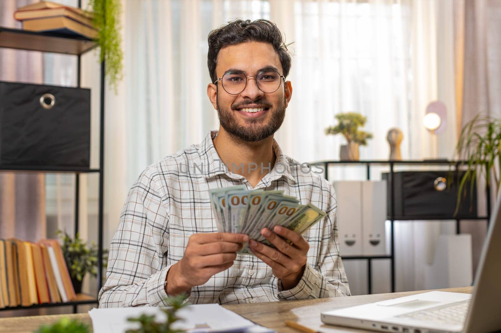 Happy young Indian businessman counting money cash and using laptop in home office. Successful excited freelancer getting bundle of money in office, calculates dividends cash money profits finances.