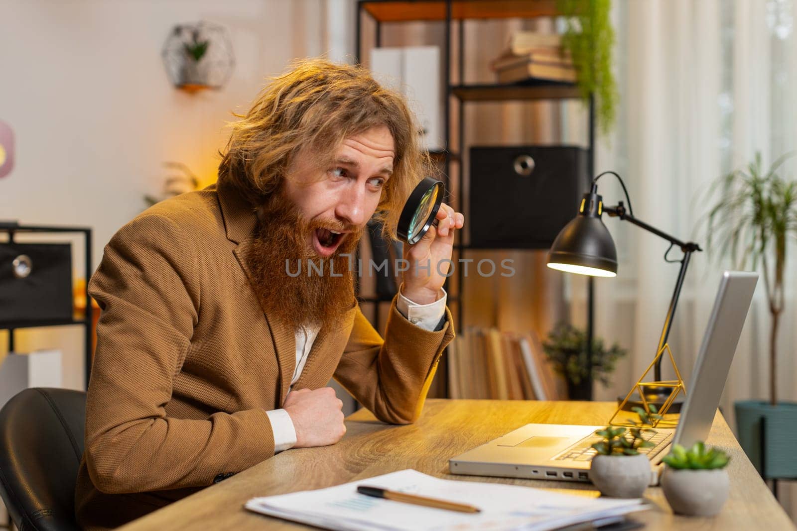 Surprised investigator researcher businessman working at home office holding magnifying glass looking at laptop screen with wow expression, searching, analyzing. Excited freelancer at home office.