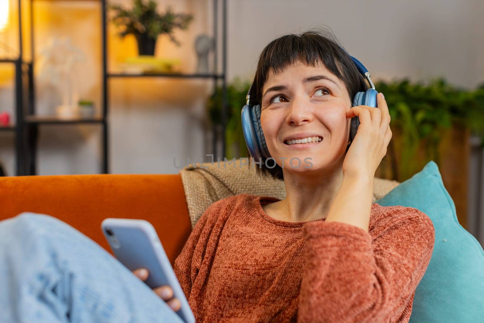 Portrait of happy girl in wireless headphones choosing listening favorite energetic dancing music on smartphone. Smiling young woman relaxing on sofa in living room at home in apartment during weekend