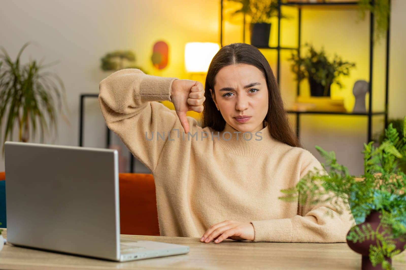 Dislike. Upset unhappy Caucasian woman showing thumb down sign gesture expressing discontent disapproval dissatisfied bad work at home office indoors. Displeased pretty young girl in room at table
