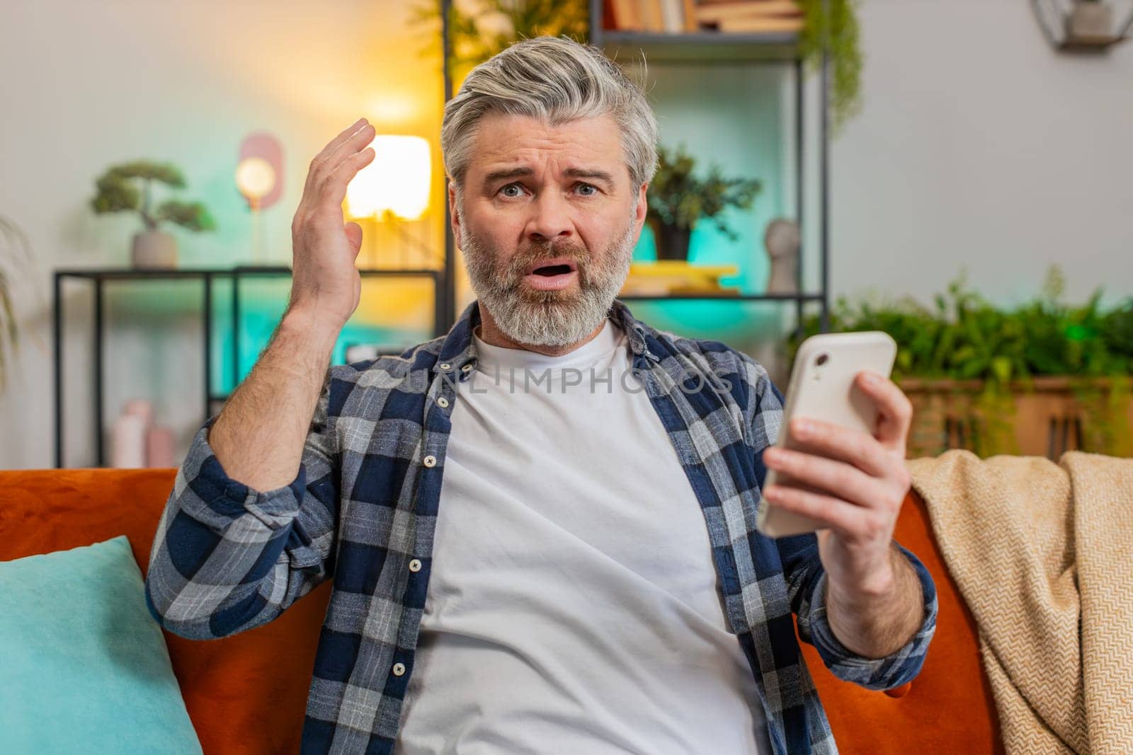 Shocked sad mature man holding smartphone reading negative message feels annoyed sitting on sofa in living room. Caucasian senior bearded old guy having gadget trouble in apartment. App crash concept.