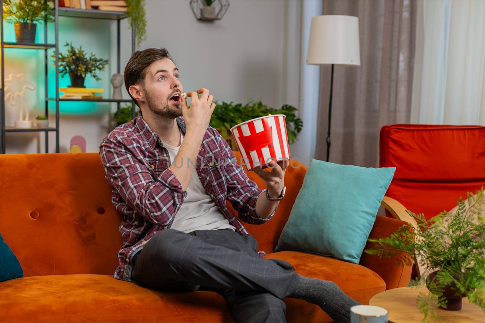 Excited man eating popcorn watching interesting tv serial sport game online social media movie content sitting on sofa in room at home. Happy Caucasian guy in casual shirt enjoying film during weekend