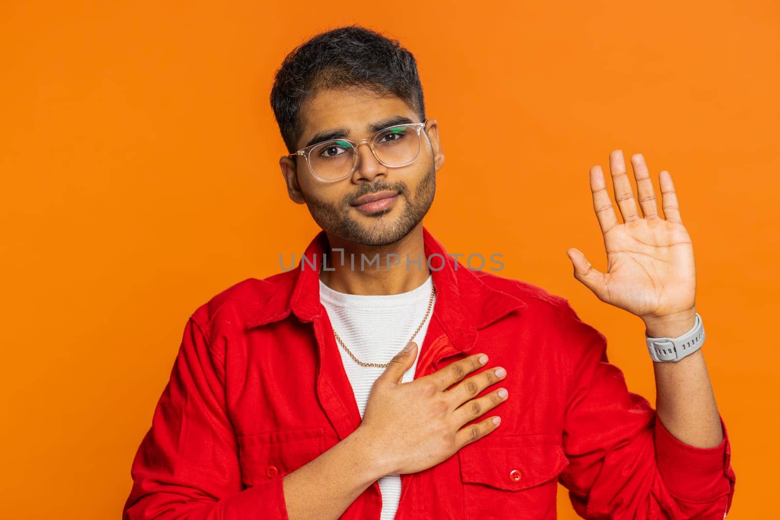 I swear to be honest. Sincere responsible Indian man raising hand to take oath, promising to be honest, to tell truth, keeping hand on chest, justice. Arabian Hindu guy isolated on orange background