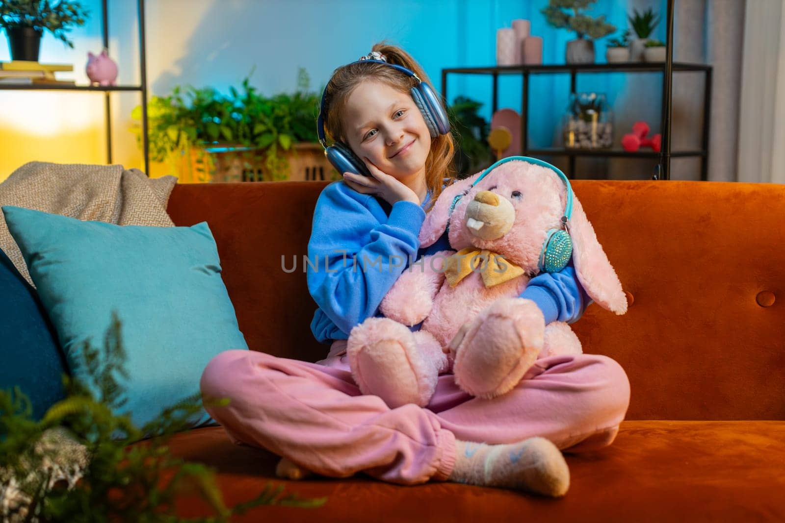 Happy Caucasian teenager girl kid in wireless headphones relaxing taking a break sits on couch at night home room listening favorite energetic dancing music. Children weekend leisure activities